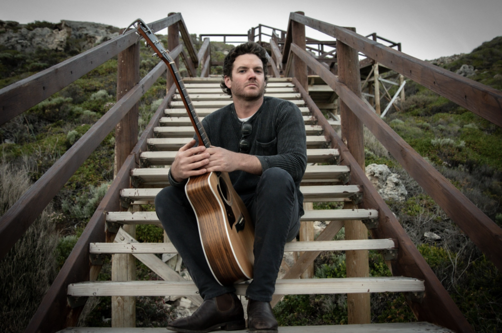 A person sits on wooden stairs outdoors, holding an acoustic guitar.